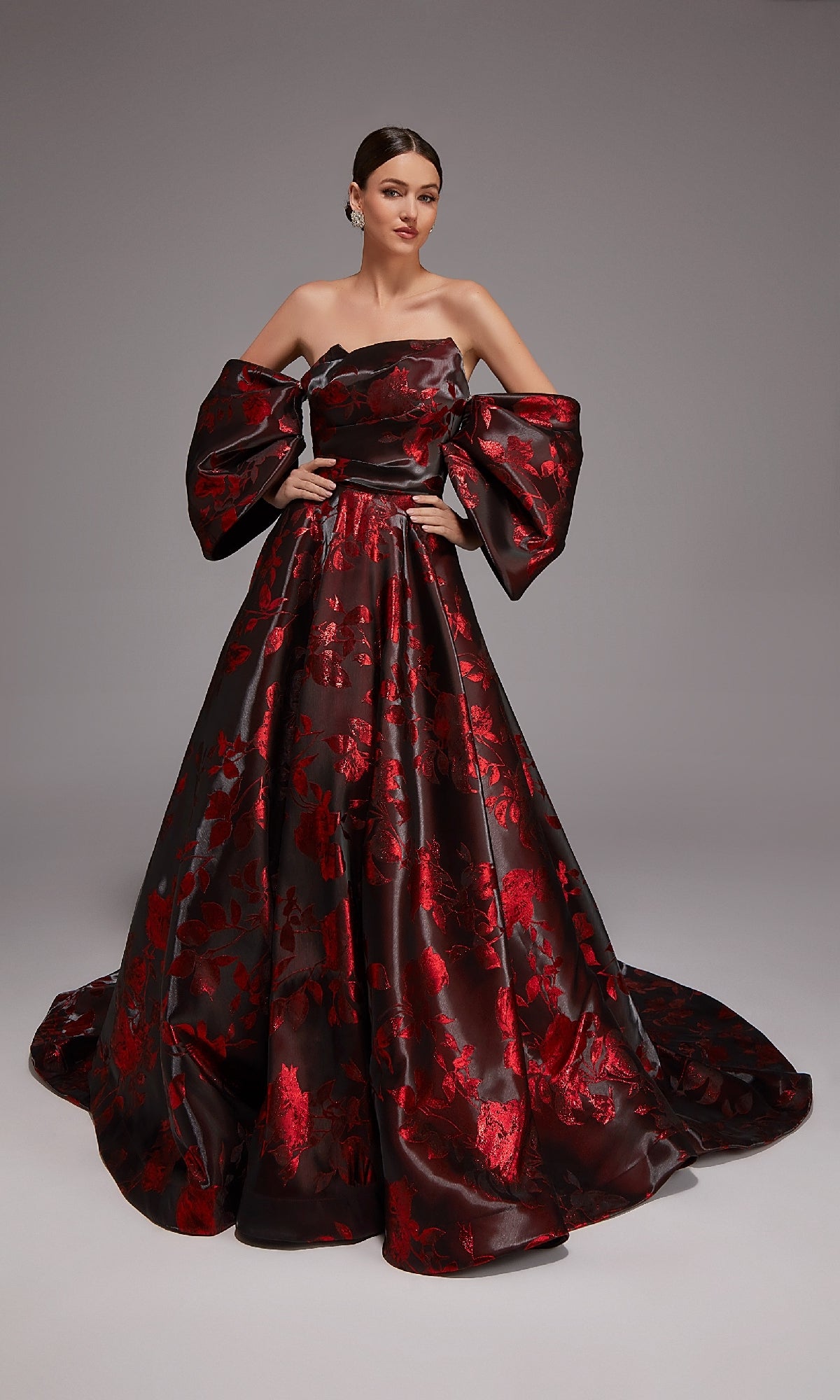 A woman stands against a plain gray background wearing the Alyce 27862 long formal dress in black/red, featuring an off-the-shoulder design, floor-length floral jacquard fabric, voluminous sleeves, and red floral patterns on a dark base.